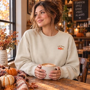Puede incluir: Una sudadera beige de cuello redondo con un pequeño bordado en el pecho izquierdo. El bordado muestra una calabaza rodeada de hojas de otoño y el texto Cosy Season en una fuente cursiva marrón. La sudadera tiene mangas largas.