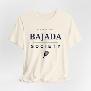 May include: A cream-coloured short-sleeve t-shirt featuring the dark blue text MEMBERS ONLY BAJADA SOCIETY and a small graphic of a paddle racket centred below the text. The shirt has a crew neckline and a standard fit with a plain back.