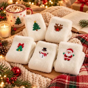 Puede incluir: Cinco pares de calcetines blancos y suaves con diseños navideños bordados, incluyendo un árbol verde, un reno marrón, una campana verde con lazo rojo, un muñeco de nieve y un Santa Claus sobre una superficie de madera.