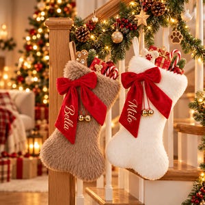 Puede incluir: Dos calcetines navideños con forma de hueso hechos de piel sintética afelpada, uno en color canela y otro en blanco. Cada uno tiene un gran lazo de terciopelo rojo con cascabeles dorados y los nombres Bella y Milo bordados.