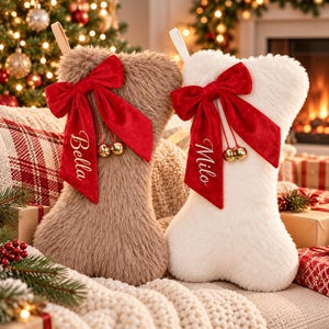 Puede incluir: Dos calcetines navideños con forma de hueso hechos de piel sintética afelpada, uno de color tostado y otro blanco. Cada uno tiene un gran lazo de terciopelo rojo con dos pequeños cascabeles dorados. El calcetín tostado tiene bordado el nombre Bella y el blanco el nombre Milo. Ambos tienen un lazo de tela para colgar.