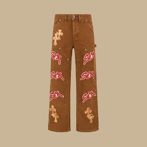 Può includere: Pantaloni in denim marrone a vita alta con chiusura a bottone. Le gambe sono decorate con diverse toppe ricamate in rosso con la scritta "sex" e applicazioni a forma di croce color beige sparse sui pannelli frontali.