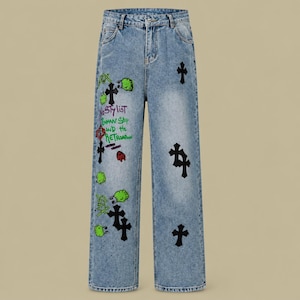 May include: Light blue acid-wash denim jeans featuring black cross appliques and colorful graphic prints. The text reads "No Stylist", "Bannan Ship", and "AND THE RETRO". The jeans have a high-waisted fit with a button and zipper closure.