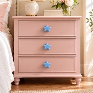 May include: A light pink wooden bedside table featuring three drawers, each fitted with a glossy, light blue star-shaped knob. The piece stands on four short, rounded feet and has a rectangular frame with clean, moulded edges on the drawer fronts.