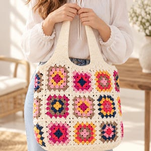 Op de afbeelding: Een gehaakte tas met een raster van kleurrijke granny squares in tinten roze, geel, blauw en bruin, omlijst door een gebroken witte rand. De tas heeft twee geïntegreerde handvatten en is gemaakt van garen in een vierkant patroon.