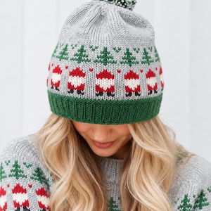 May include: A grey knit beanie featuring a green ribbed brim and a pom-pom on top. The hat displays a repeating pattern of green pine trees and red and white Santa Claus figures, with small red hearts interspersed between the festive motifs.