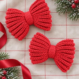 May include: Two bright red knitted bows featuring a ribbed texture and a central knot detail. The bows are arranged on a light-coloured fabric with a subtle grid pattern, accompanied by green pine sprigs and red berries.