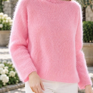 May include: A fuzzy, light pink long-sleeve jumper with a wide boat neckline. The knit garment features a soft, brushed texture and long sleeves, paired with plain white trousers. The jumper has a relaxed fit and a solid pastel pink colour.