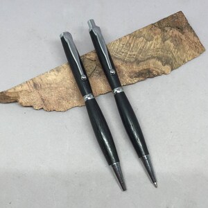 Könnte beinhalten: Zwei schwarze Holzstift mit silbernen Akzenten. Die Stifte liegen auf einem Stück Holz mit einer natürlichen Maserung.