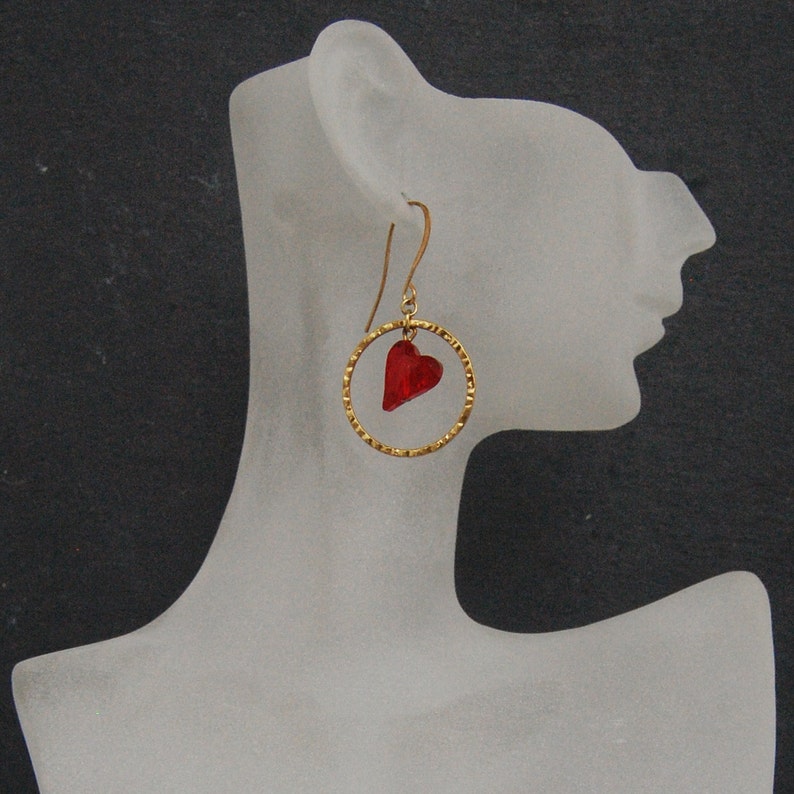 May include: Gold hoop earrings with a red heart charm. The earrings are on a white mannequin head.