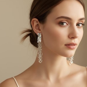 May include: Silver dangle earrings. Each earring features a textured rectangular top with a cascade of small, square-shaped charms. The earrings are designed to hang down, creating a long, elegant look. The earrings are made of a shiny, metallic material.