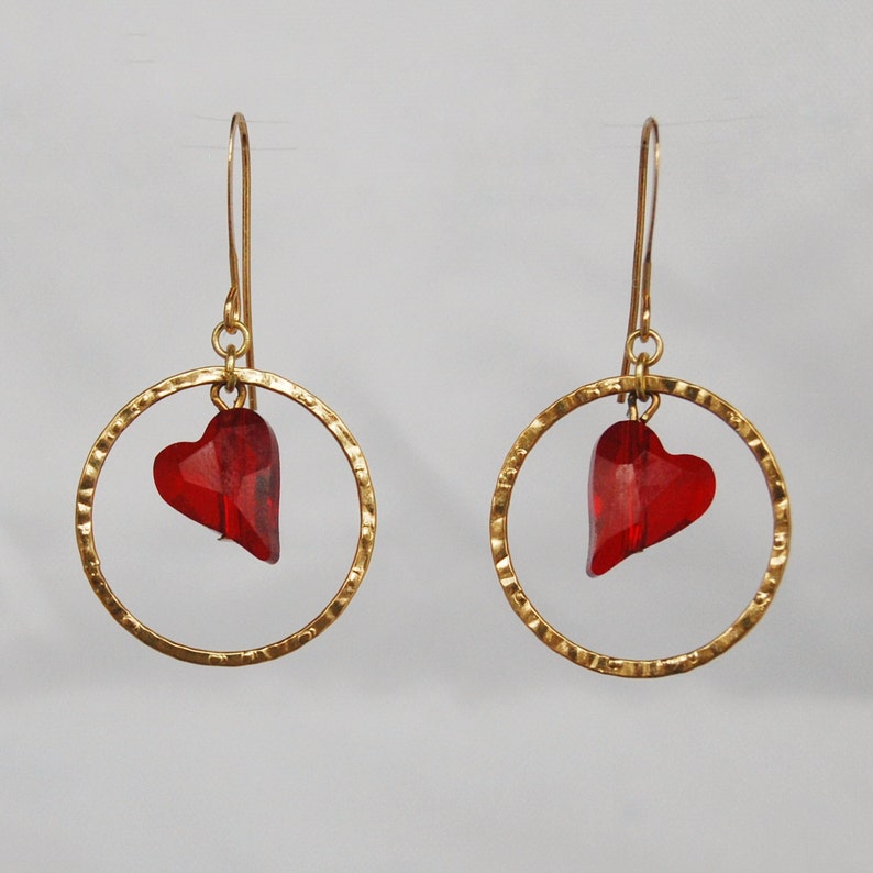 May include: A pair of gold hoop earrings with a red heart charm dangling from each hoop.