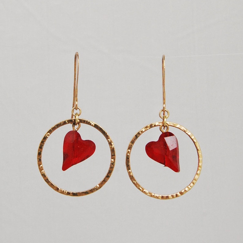 May include: Gold hoop earrings with a dangling red heart charm. The hoops are hammered for a textured look.