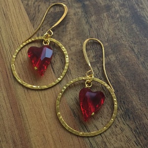 May include: A pair of gold hoop earrings with a red heart-shaped crystal dangling from each hoop.