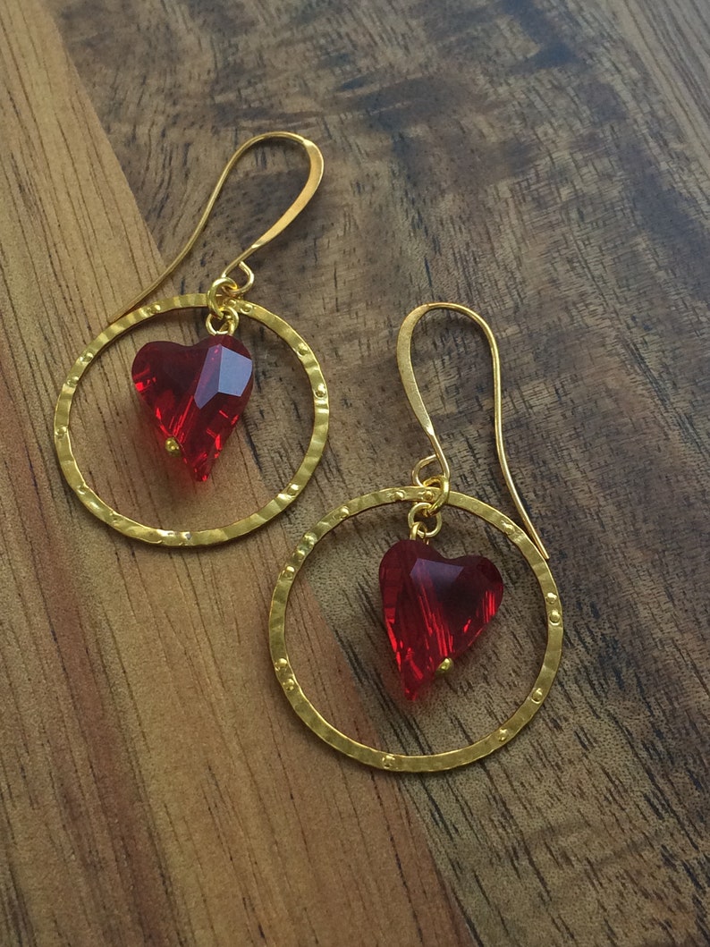 May include: A pair of gold hoop earrings with a red heart-shaped crystal dangling from each hoop.