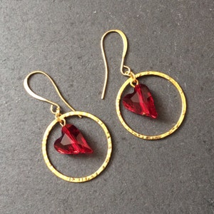 May include: A pair of gold hoop earrings with a red heart-shaped crystal dangling from each hoop.