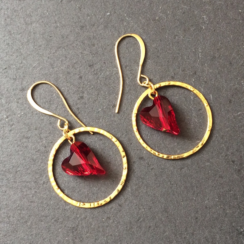 May include: A pair of gold hoop earrings with a red heart-shaped crystal dangling from each hoop.