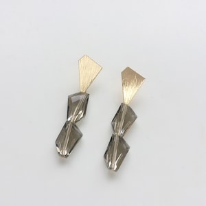 May include: A pair of gold-coloured earrings with a geometric design. Each earring features a textured, triangular top and two smoky grey, faceted crystal-like shapes suspended below. The earrings are set against a white background.