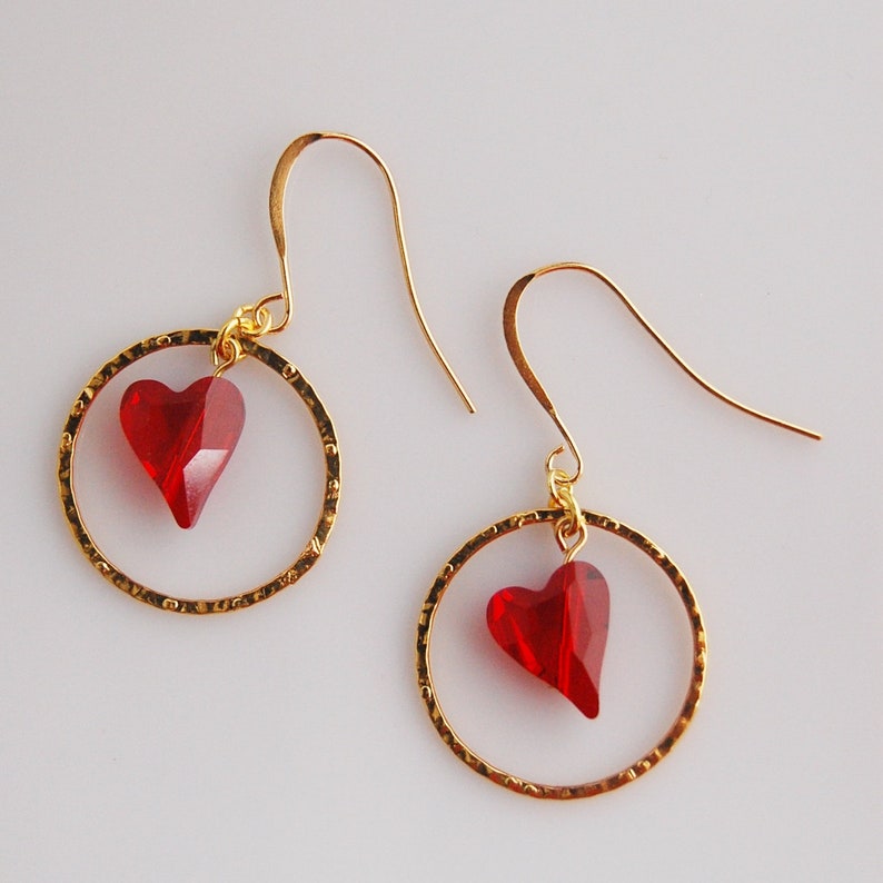 May include: A pair of gold hoop earrings with a red heart-shaped crystal charm hanging from each hoop.