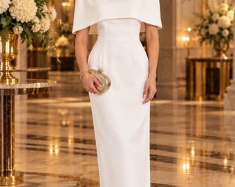 Op de afbeelding: Een vloerlange witte jurk met een hoge halslijn en een geïntegreerde cape die de schouders bedekt. Het kledingstuk wordt gecombineerd met metallic gouden pumps met een puntige neus en een kleine, gestructureerde gouden clutch.