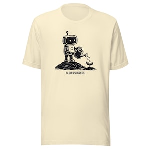 May include: A cream-colored t-shirt featuring a black graphic of a small robot watering a tiny plant with a watering can. The robot stands on a mound of dirt, and the text SLOW PROGRESS. is printed in black capital letters below the illustration.