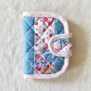 May include: A quilted fabric card holder featuring a blue diamond-stitched pattern, a central panel with strawberry and elephant prints, and a light pink gingham border. The accessory is secured with a single light pink plastic snap button.