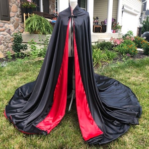 Op de afbeelding: Een zwarte satijnen cape met een levendige rode voering en bies wordt getoond op een mannequin. De cape heeft een hoge kraag en een striksluiting bij de hals. Het lange, vloeiende ontwerp is uitgespreid op een grasachtig oppervlak.