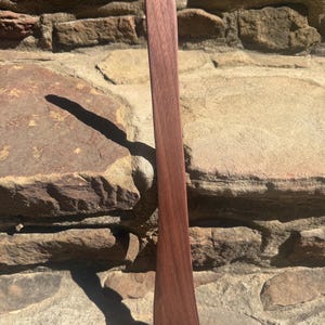 May include: A long, slender wooden kitchen spatula with a dark brown, natural wood grain finish. The utensil features a flat, slightly curved blade at the bottom and a rounded handle at the top, showcasing a smooth, polished surface texture.
