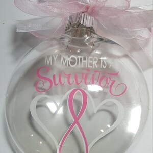 May include: Clear glass Christmas ornament with a pink ribbon and white lettering that reads "My Mother is a Survivor".