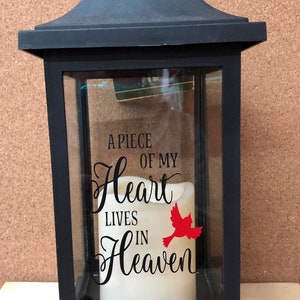 May include: Black lantern with a glass panel featuring a white candle and a red cardinal silhouette. The glass panel has the text "A piece of my heart lives in heaven".