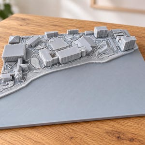 May include: A gray 3D printed architectural model featuring a detailed city layout with various buildings, structures, and pathways set against a flat, smooth base representing a waterfront area, capturing a miniature urban landscape.