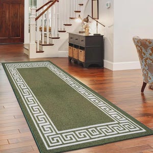 May include: A rectangular runner rug featuring a solid olive green centre and a wide white border decorated with a repeating geometric Greek key pattern. The rug is positioned lengthwise on a polished wooden floor, showcasing its flat-woven texture.