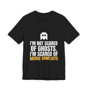 May include: A black short-sleeved t-shirt featuring a white pixelated ghost icon above the white text "I'M NOT SCARED OF GHOSTS. I'M SCARED OF" and the yellow text "MERGE CONFLICTS" printed in a bold, sans-serif font on the centre of the chest.