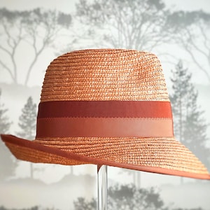 以下が含まれることがあります： 幅広の錆色のバンドが付いたストローフェドラハット。帽子は、わずかに湾曲したつばを持つクラシックな形状をしています。帽子は森のシーンを背景に表示されています。