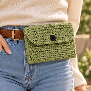 May include: A sage green crochet bum bag featuring a rectangular shape, a fold-over flap, and a single black circular button closure. The bag is constructed with a dense, textured stitch pattern and is worn attached to a brown leather belt.