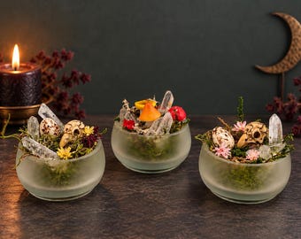 May include: Three small, round, frosted glass bowls filled with green moss, clear quartz crystals, small bird skulls, and colorful miniature flowers. The arrangements are displayed on a dark surface with a lit candle and moon decor.