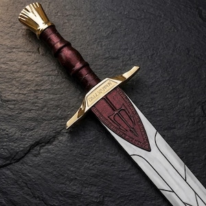May include: A sword with a metallic silver blade featuring dark etched lines. The hilt has a deep red grip, a gold-colored pommel, and a gold crossguard engraved with text. A red triangular emblem with a trident symbol sits on the blade.