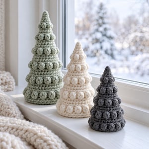May include: Three cone-shaped crochet trees in sage green, cream, and charcoal gray. Each tree features a textured bobble stitch pattern arranged in horizontal rows, creating a dimensional surface. The trees are arranged in a line.