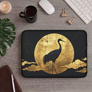 Könnte beinhalten: Eine schwarze Laptoptasche mit einem großen, kreisförmigen, goldfarbenen Mond mit rissiger Textur. Ein schwarzer Kranich steht in der Mitte des Mondes, umgeben von stilisierten goldenen Wolken auf einem schwarzen Hintergrund mit feiner Marmorierung.