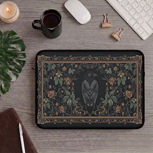 May include: A rectangular black laptop sleeve featuring a central illustration of a crow surrounded by intricate floral patterns and foliage in shades of muted green and copper, all enclosed within a decorative, ornate rectangular border frame.