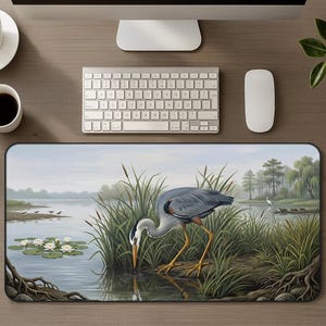 May include: A rectangular desk mat featuring a detailed illustration of a grey heron standing in shallow water among tall green reeds and cattails. The scene includes white water lilies on the left and a distant shoreline with trees under a pale sky.
