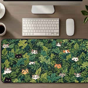 Puede incluir: Alfombrilla de ratón rectangular con un diseño de jungla verde y numerosas caras de gatos asomando entre las hojas. Los gatos tienen diferentes patrones y colores de pelaje, añadiendo un toque lúdico al accesorio de escritorio.