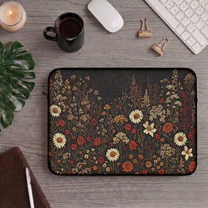 May include: A rectangular laptop sleeve featuring a dark background with a dense, colorful pattern of wildflowers in shades of red, orange, yellow, and white. The sleeve has a black zipper closure along the top edge and a smooth, flat surface.