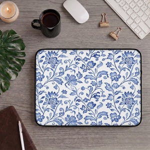 Puede incluir: Una funda rectangular para portátil con fondo blanco y un patrón floral azul repetitivo de flores, hojas y enredaderas estilizadas. La funda tiene un borde negro y un cierre de cremallera a lo largo del borde superior para proteger el equipo.
