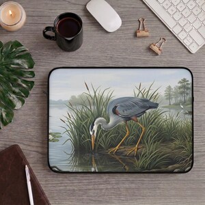 May include: A rectangular laptop sleeve featuring a detailed illustration of a grey heron standing in shallow water among tall green reeds and cattails. The bird has a long neck, a sharp yellow beak, and slender yellow legs, with its reflection visible in the water.