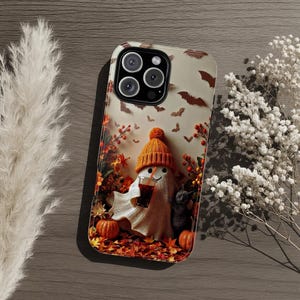 May include: A smartphone case featuring a graphic of a white ghost wearing an orange knit beanie, holding a dark beverage with a straw, accompanied by a small black cat, orange pumpkins, autumn leaves, and brown bats on a light background.