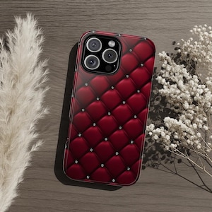 May include: A burgundy phone case with a quilted pattern and small, sparkling embellishments. The case has a glossy finish and a dark border, designed to fit a smartphone. The case is shown on a wooden surface.