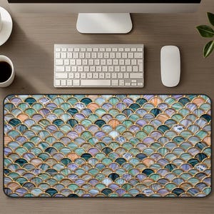 May include: A rectangular mouse pad with a mermaid scale design. The pattern features overlapping, fan-shaped elements in shades of blue, green, purple, and peach, outlined with gold. The mouse pad is on a wooden desk with a keyboard, mouse, and coffee cup.