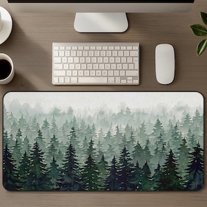 May include: A rectangular desk mat featuring a watercolor-style illustration of a dense forest with layers of evergreen trees in varying shades of dark green and sage, fading into a light, misty gray background at the top edge.