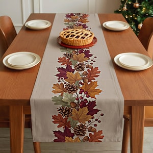 May include: A light gray table runner featuring a central vertical print of autumn leaves in shades of orange, red, and brown, interspersed with pinecones, acorns, and red berries. A lattice-crust pie sits on a red plate on the runner.